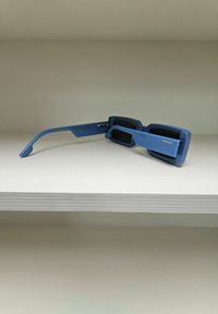 Lunettes de soleil rectangulaires bleu avec des montures épaisses, dotées de verres foncés et d'une finition mate lisse. Le nom de la marque "KOMONO" est visible sur le côté.