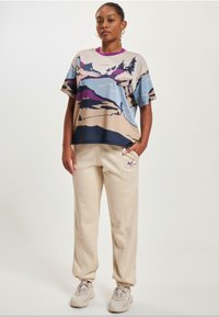 Grafisches T-Shirt mit kurzen Ärmeln, das eine Berglandschaft in Beigetönen, Blau und Violett zeigt. Kombiniert mit beigen Jogginghosen und hellen Sneakern.