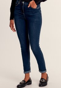 Donkerblauwe, high-waisted jeans met een slank silhouet, opgerolde mouwen en subtiele stiksels. Draag ze met zwarte loafers.