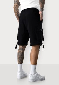 Schwarze Cargo-Shorts mit elastischem Bund, ausgestattet mit seitlichen Taschen und verstellbaren Bindebändern am Saum, getragen mit weißen Sneakers und Socken.