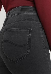 Jeans de denim negro con un diseño ajustado, costuras sutiles y una etiqueta con el nombre de la marca en la parte trasera. Incluye un bolsillo trasero curvado.