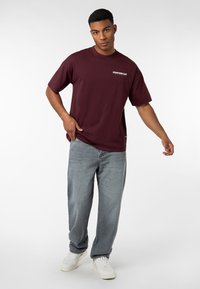 Burgunder Baumwoll-T-Shirt mit kurzen Ärmeln und weißem "DOWNTOWN CLUB"-Text. Kombiniert mit hellgrauen Denim-Jeans und weißen Sneakers.