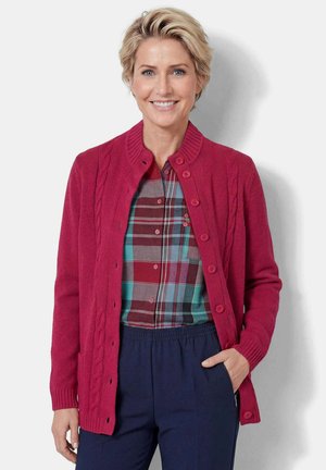 Vrouw draagt een rode gebreide cardigan met knopen over een meerkleurig geruit overhemd en donkerblauwe broek, glimlacht met hand in zak tegen een witte achtergrond.