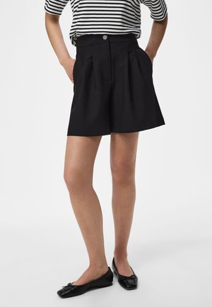 Person iført sorte højtaljede plisserede shorts, sorte balletsko og en sort-hvid stribet kortærmet top, stående med hænderne i lommerne.