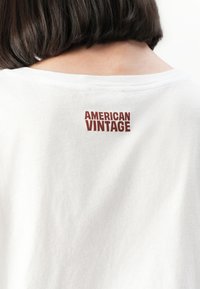 Person med en hvit T-skjorte med "AMERICAN VINTAGE" trykket i rødt på øverste del av ryggen, mørkt hår synlig bakfra.