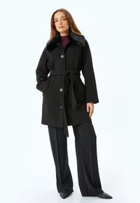 Cappotto di lana nera con colletto in pelliccia sintetica, chiusura con bottoni, cintura rimovibile, e maniche lunghe, abbinato a pantaloni neri a gamba larga.