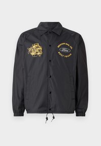 Black windbreaker jacket with ribbed cuffs, snap buttons, and drawstring hem. Features yellow graphic prints of an engine and brand logos.