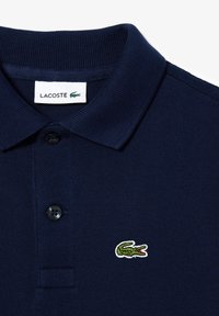 Tummansininen puuvillapaita, jossa on ribbireunus, kaksi mustaa nappia ja brodeerattu vihreä krokotiili-logo vasemmassa rinnassa.