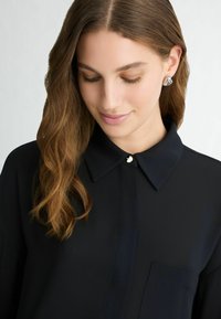 Navy blouse with a pointed collar, single button closure, and a front pocket. The fabric has a smooth texture with subtle sheen.