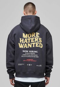 Person wearing a dark hoodie with text "MORE HATERS WANTED," and smaller text humorously advertising "Now hiring" with a fictional phone number.