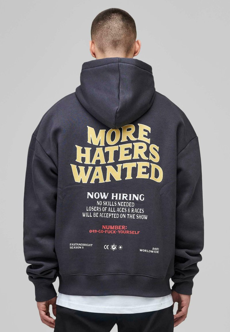 Person wearing a dark hoodie with text "MORE HATERS WANTED," and smaller text humorously advertising "Now hiring" with a fictional phone number.