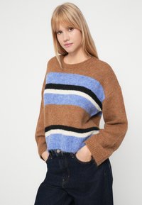 Ruda megzta palaidinė, papuošta horizontaliomis juostomis šviesiai mėlynos, juodos ir baltos spalvos. Laidūs siluetas su nuleistais pečiais ir apvaliu kaklu.