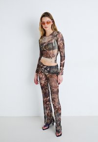 Langarm, aansluitend topje en uitlopende broek in een bruine tie-dye print. Gestructureerde sheer stof met gelaagde details; gecombineerd met getinte zonnebril.