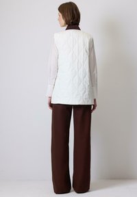 Hvid quiltet vest med ærmeløst design, sammen med brune brede bukser. Vestens struktur har et tekstureret mønster og en lige pasform.