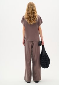 Top ampio di un tenue color malva e pantaloni a gamba larga realizzati in un tessuto morbido e fluido. Borsa nera trapuntata con tracolla tenuta in mano.