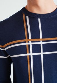Navy Pullover mit horizontalen und vertikalen Streifen in Weiß und Senf. Verfügt über einen gerippten Halsausschnitt und ein weiches, strukturiertes Strickmaterial.