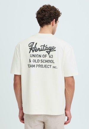 Homme aux cheveux bouclés portant un t-shirt écru avec un texte noir "Heritage Union of '63 & Old School Team Project Inc." au dos, debout devant un fond pâle.