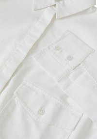 Camicia bianca con bottone realizzata in cotone strutturato, con colletto classico e due tasche frontali con chiusura a bottone.
