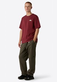 Maroon short-sleeve t-shirt featuring a front logo, paired with dark green pants with zippers, worn with black sneakers.