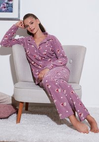 Violetiniai flisiniai pižamos su gėlių raštu, su apykakle, mygtukais ir griovelių tekstūra. Modelis sėdi ant šviesiai pilkos kėdės.