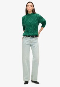 Pull en maille torsadée verte avec un col montant, associé à un jean large bleu clair et des bottines noires. Design simple et texturé.