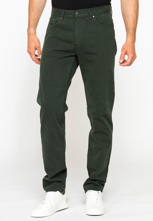 Uomo che indossa pantaloni casual verde scuro e sneakers bianche, in piedi davanti a uno sfondo bianco semplice.