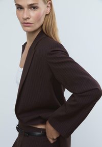 Blazer marron à rayures, avec des revers marqués et des manches longues, associé à un haut blanc, présentant une silhouette ajustée et une texture lisse.