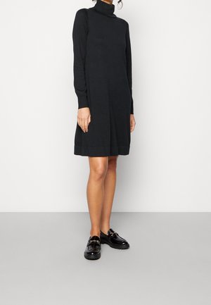 Kobieta w czarnej, długorękawowej, dzianinowej sukience z golfem oraz czarnych błyszczących mokasynach, stojąca na tle jednolitego, jasnego tła.