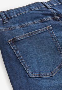 Tumma sininen farkut, joissa näkyy takatasku, korostettuna oranssilla ompeleella saumoissa. Sujuva rakenne ja vähäinen kiilto.