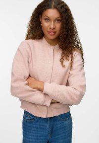 Mujer con pelo rizado, vistiendo una chaqueta abotonada de un rosa claro y vaqueros azules, de pie con los brazos cruzados contra un fondo blanco.