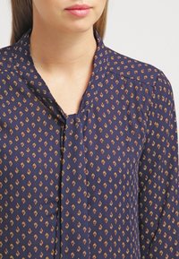 Donna che indossa una blusa blu navy con un piccolo motivo floreale arancione e giallo e dettagli con scollatura a chiave.