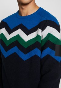 Sweater featuring a zigzag pattern in blue, green, white, and navy, with a round neckline and textured knit. Black accent on the shoulder.
