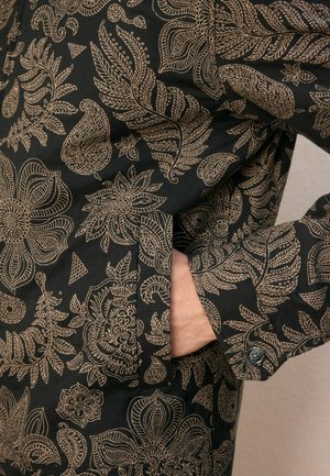 Schwarzer Stoff mit kunstvollen hellbraunen Blumen- und Blättermustern, der eine in eine Seitentasche eines geknöpften Ärmels geschobene Hand zeigt.