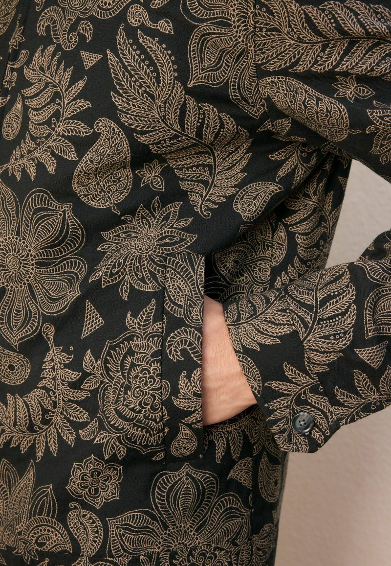 Tissu noir avec des motifs floraux et de feuilles beige complexes, montrant une main glissée dans une poche latérale d'un vêtement à manche boutonnée.