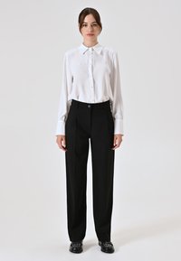 Camicia bianca con colletto, abbinata a pantaloni neri a vita alta. I pantaloni presentano un design plissettato e sono sagomati. Scarpe nere completano il look.