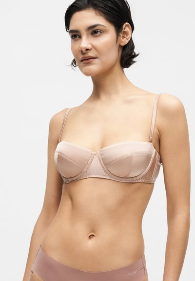 Frau, die einen hautfarbenen trägerlosen BH mit dünnen verstellbaren Trägern und fliederfarbige nahtlose Unterwäsche trägt, vor einem schlichten hellen Hintergrund stehend.