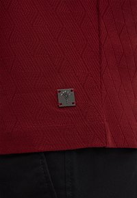 Maglione rosso a maglia con un motivo geometrico testurizzato e un'etichetta metallica sul fondo, abbinato a pantaloni scuri.