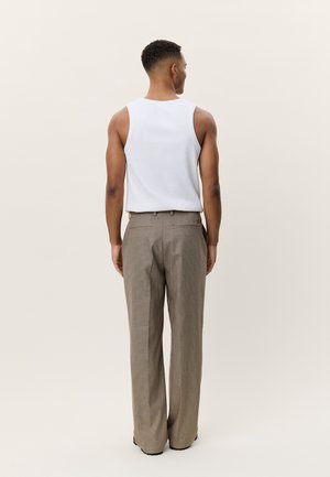 Homme vu de dos portant un débardeur blanc et un pantalon large à carreaux beige sur un fond clair uni.