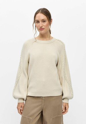 Femme aux cheveux attachés portant un pull en maille crème avec des détails tressés sur des manches volumineuses et un pantalon marron clair sur un fond uni.