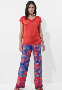 Rotes Satinoberteil mit kurzen Ärmeln und V-Ausschnitt, kombiniert mit blauen weit geschnittenen Hosen im Blumenmuster mit roten Akzenten und detaillierten Verzierungen.