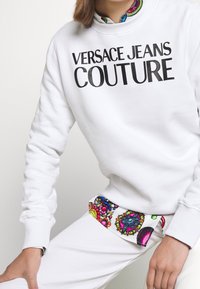 Bílá mikina s logem "VERSACE JEANS COUTURE" v černé barvě, vystavená s barevným páskem zdůrazňujícím bílé kalhoty.