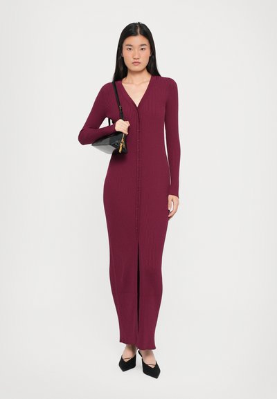 Long, burgundy ribbed dress with a V-neck, full-length, front button closure, and slit at the bottom; styled with a black handbag and pointed heels.