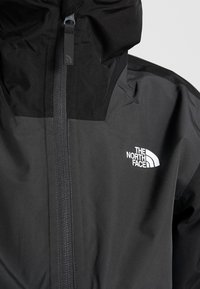 Schwarze wasserdichte Jacke mit hohem Kragen, Reißverschluss und weißem Logo von "The North Face" auf der linken Brust. Glatte Stofftextur.