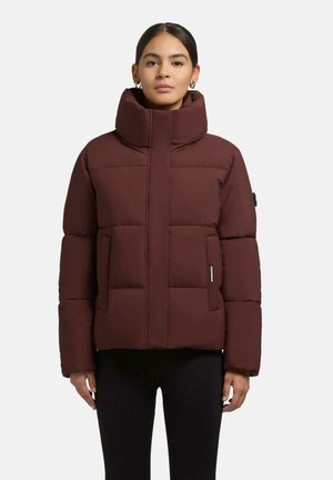 Donna che indossa un giubbotto invernale imbottito di un rosso scuro con collo alto e pantaloni neri, in piedi davanti a uno sfondo chiaro e uniforme.