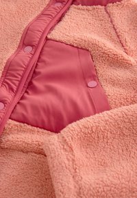 Růžová fleecová bunda s měkkou texturovanou látkou a hladou vnitřní podšívkou, s patentkami podél předního otvoru a kapsy.