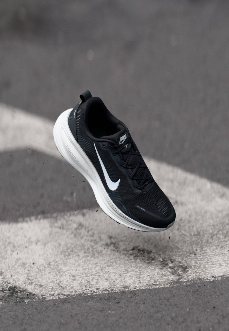 Gheată de alergare neagră cu un superior din plasă, swoosh-ul Nike alb și o talpă albă cu amortizare. Designul include o bandă pentru a fi trasă și modele texturate.