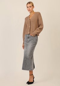 Cardigan lavorato a maglia beige con trama a coste, dotato di scollo a V e tre bottoni, abbinato a una gonna di jeans grigia con dettagli borchiati.