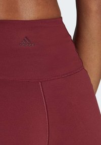 Burgundowe legginsy o gładkiej teksturze, z wysokim stanem i subtelnym wytłaczanym logo Adidas z boku.