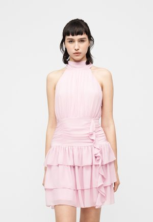 Giovane donna con capelli corti e scuri che indossa un vestito senza maniche color rosa chiaro con balze a più strati e vita arricciata.