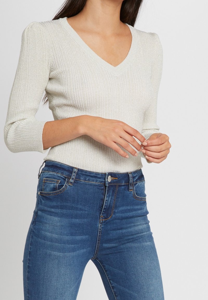 Femme portant un pull léger à nervures avec un col en V et des épaules bouffantes, ainsi qu'un jean bleu taille haute, les mains doucement croisées devant elle.